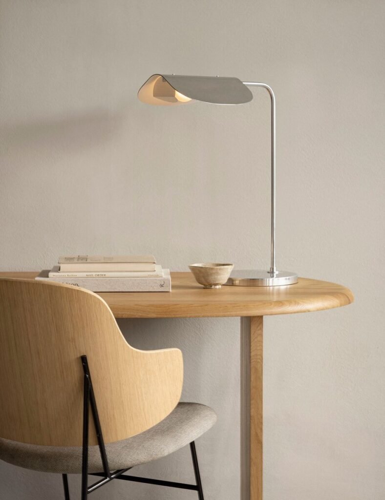 Minimalist Scandinavian-style wooden desk setup with a modern metal desk lamp, a curved wooden chair with fabric seat, a small ceramic bowl, and stacked neutral-toned books against a light beige wall.
